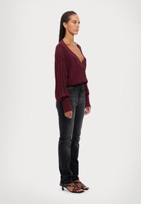 Suéter burdeos de punto trenzado con escote en V profundo, combinado con jeans negros ajustados y tacones negros de tiras. Textura suave y diseño contemporáneo.
