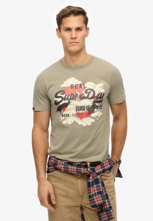 Camiseta de algodón verde olivo con un gran estampado gráfico de nubes y texto en negro y rojo. Combinada con una camisa de franela a cuadros atada a la cintura.