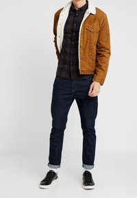 Bruine corduroy jas met een witte fleece kraag, gedragen over een donker geruit overhemd, gecombineerd met donkere spijkerbroek en zwarte sneakers.