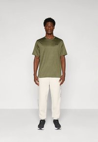 Hombre de pie mirando al frente, vistiendo una camisa de manga corta color verde oliva, pantalones blancos y zapatos deportivos negros sobre un fondo claro y liso.