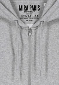 Felpa grigia con zip realizzata in cotone biologico. Presenta una zip argento, stringhe regolabili e un'etichetta stampata con le istruzioni per la cura.