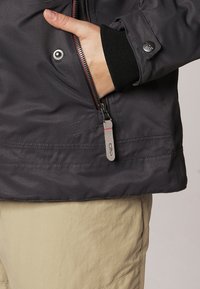 Main partiellement dans la poche zippée d'une veste gris foncé avec une tirette de fermeture éclair visible au logo CMP, pantalon beige en dessous.