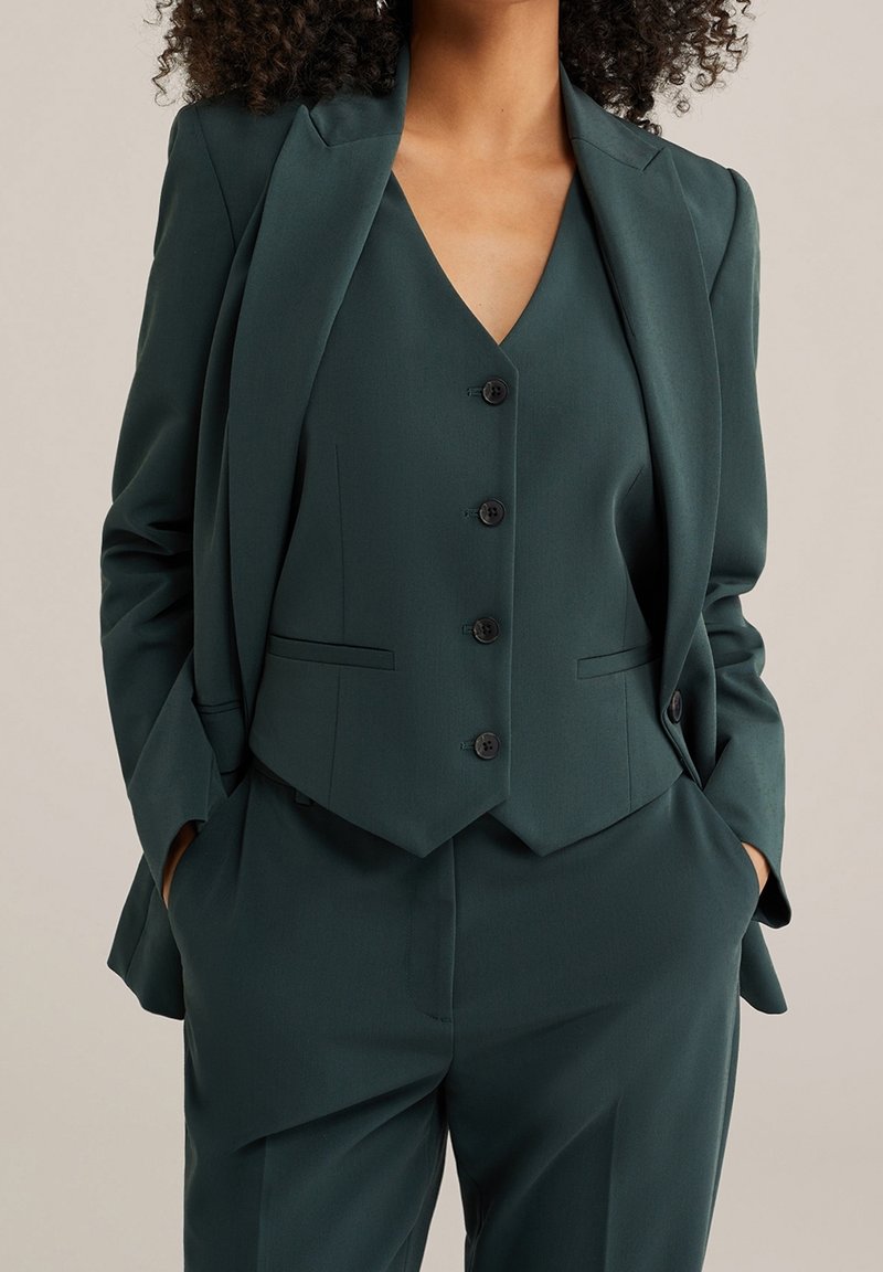 Femme portant un costume sur mesure vert foncé avec un gilet boutonné, une veste et un pantalon, les mains dans les poches de manière décontractée, sur un fond neutre.