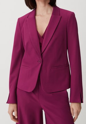 Femme portant une veste de tailleur magenta avec un bouton et un pantalon assorti, debout, les bras détendus le long du corps.