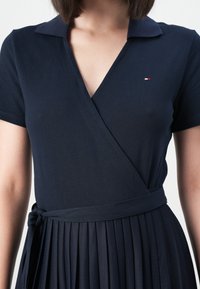 Tummansininen kietaisumekko, jossa on kauluksellinen pääntie, lyhyet hihat ja pleatattu helma. Mukana on vyötärölle sidottu nauha ja hienovarainen logoyksityiskohta.