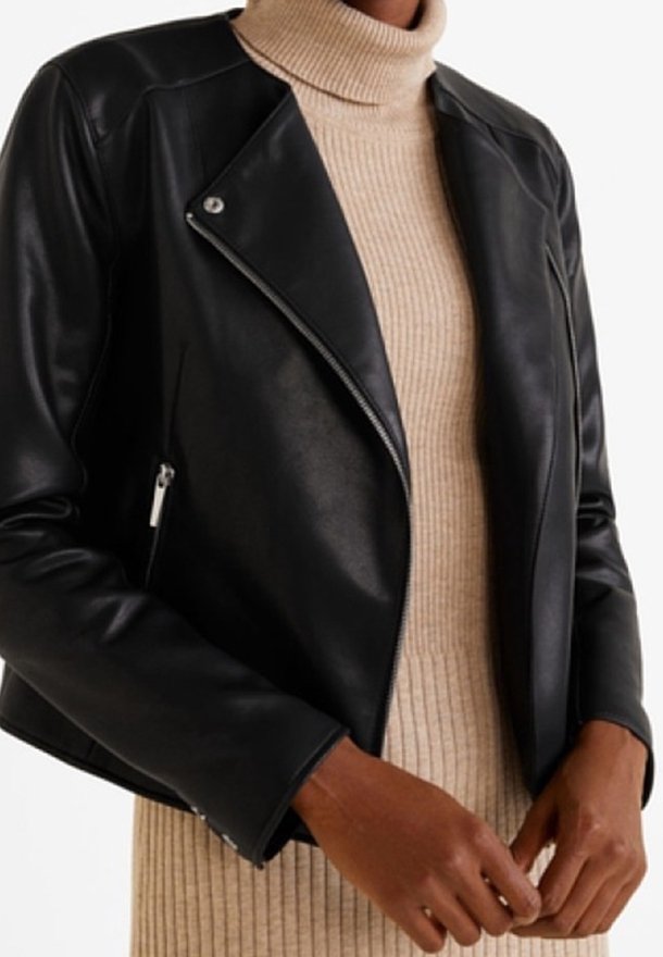 Giacca da moto in pelle nera con zip diagonale, design aderente e texture liscia, indossata sopra un dolcevita beige a coste.