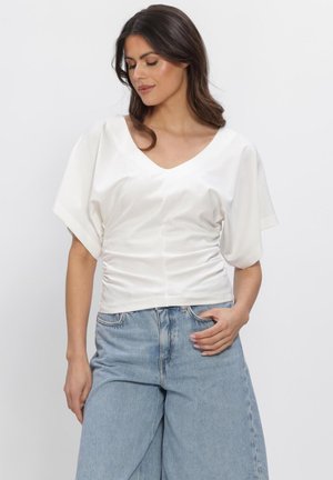 Frau mit braunen Haaren trägt eine weiße, geraffte Bluse mit V-Ausschnitt, weiten Ärmeln und hellblauen Jeans mit hoher Taille, posiert mit einer Hand in der Tasche.