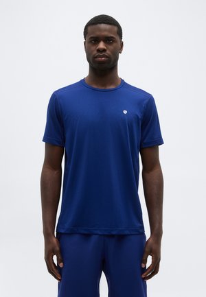 Άνδρας που στέκεται με το βλέμμα προς τα εμπρός, φορώντας απλό μπλε αθλητικό πουκάμισο και κοντό παντελόνι που ταιριάζουν, σε λευκό φόντο.