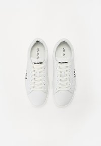 Bílé nízké tenisky Valentino s bílými tkaničkami a černým brandingem na jazyku a bocích, pohled shora na bílém pozadí.