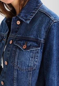 Primer plano de una persona que lleva una chaqueta de mezclilla azul con botones de cobre y costuras naranjas visibles, mostrando el cuello y el bolsillo del pecho.