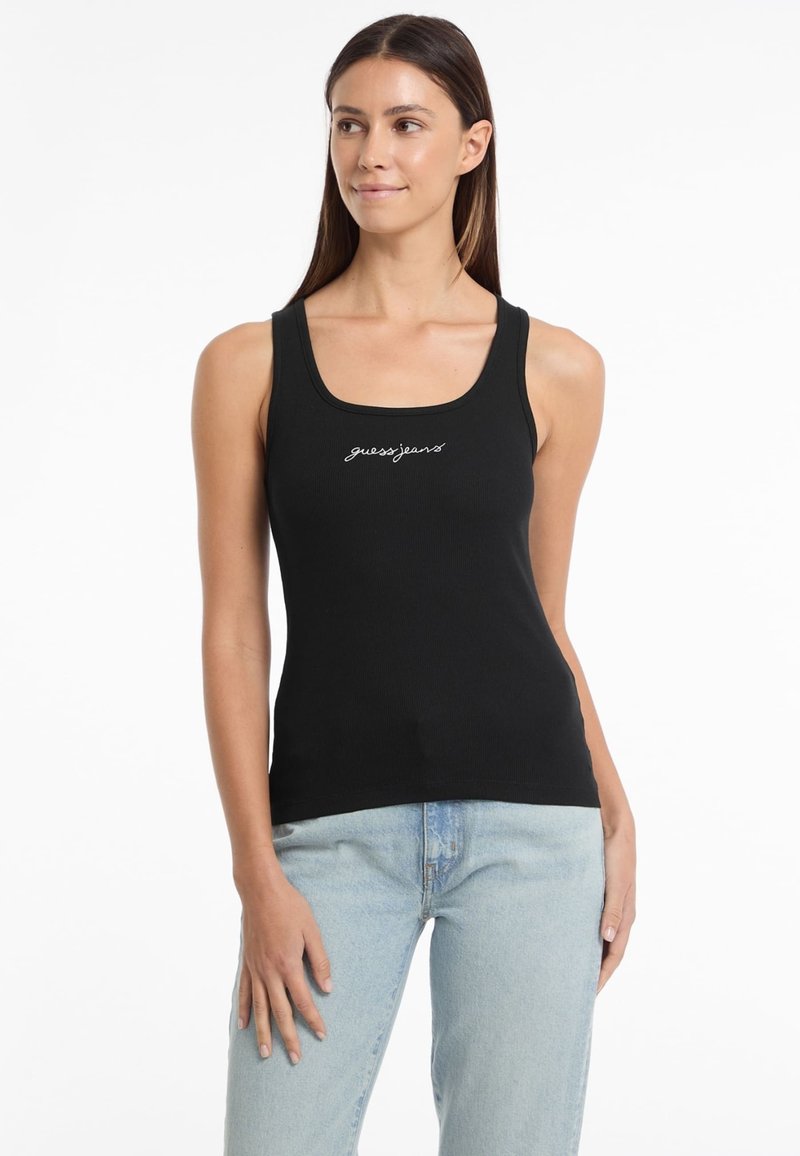 Black ribbed tank top featuring a square neckline and "guessjeans" logo in white embroidery, paired with light blue denim jeans.