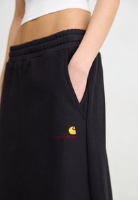 Crne jogger hlače od mekog materijala s rebrastim pojasom, bočnim džepovima i žutom i crvenom Carhartt logotipom sprijeda.
