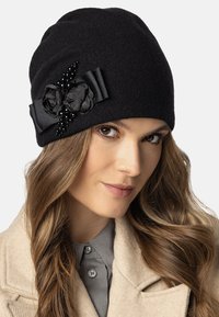 Gorro de fieltro negro con una cinta de satén y dos flores de tela decoradas con pequeñas perlas. Textura suave y diseño ajustado.