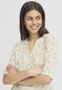 Lichte beige blouse met korte pofmouwen, versierd met een patroon van kleine roze watermeloenschijfjes. Knopdetail bij de halslijn.