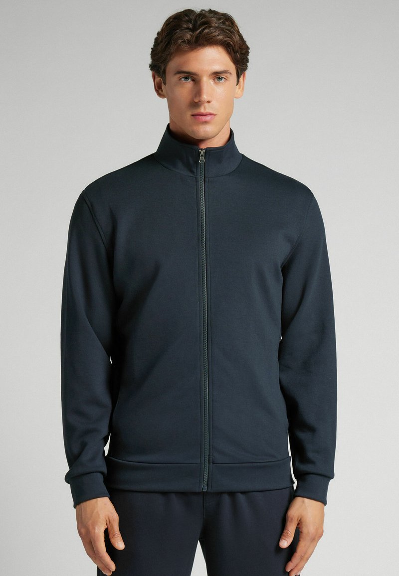 Chaqueta navy con cremallera y alto cuello, puños y cinturilla de canalé. Tejido suave con un diseño minimalista y un sutil detalle de costura.