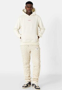 Crème hoodie met rode tekst, gecombineerd met crèmekleurige broek met een textuur en een gevlochten patroon. Zwarte en beige sneakers maken de outfit compleet.