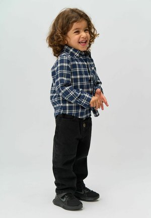 Camisa de franela a cuadros en azul marino y blanco con un bolsillo, pantalones de mezclilla negros y deportivas negras. El niño tiene el pelo rizado y está de pie con las manos entrelazadas.