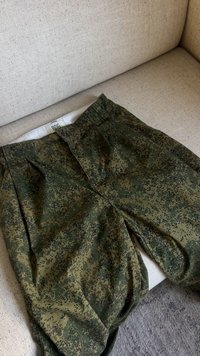 Calças de camuflagem digital verdes dobradas num sofá bege com um tapete cinza visível ao lado.