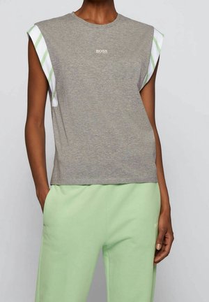 Femme portant un haut sans manches gris BOSS avec un détail d'épaule rayé blanc et vert, et un pantalon vert clair, main dans la poche.