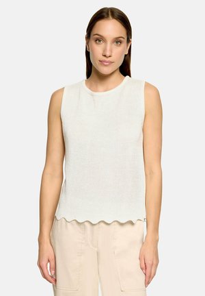 Vrouw met steil bruin haar die een mouwloos wit gebreid topje met geschulpte zoom en lichtbeige broek draagt, staand tegen een effen achtergrond.