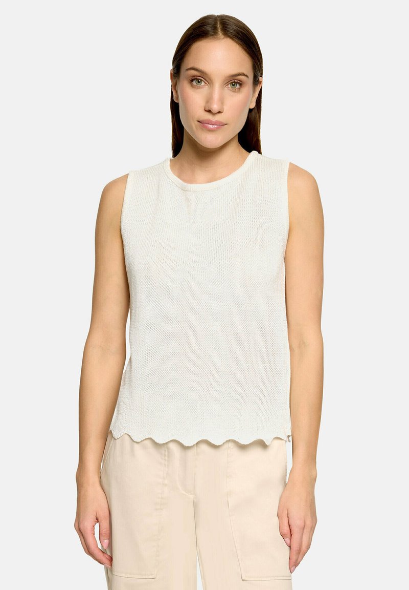 Femme aux cheveux bruns et raides portant un haut blanc en maille sans manches avec un ourlet festonné et un pantalon beige clair, debout sur un fond uni.