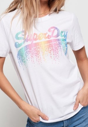Femme portant un T-shirt blanc avec le texte "SuperDry" coloré en pixels et un jean bleu, mains dans les poches de manière décontractée, fond clair.