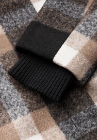 Närbild av en rutig ullärm med beige, grå, svart och vita rutor, som har en svart ribbad mudd.