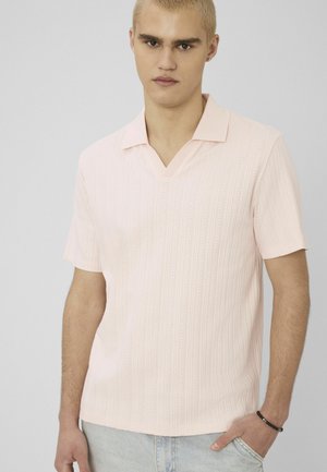 Jeune homme aux cheveux blonds courts portant un polo à manches courtes texturé rose pâle et un jean bleu clair, debout avec une main dans la poche.