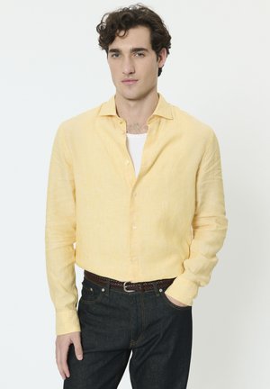 Svetložltá ľanová košeľa na gombíky s dlhými rukávmi a uvoľneným golierom, doplnená tmavohnedými džínsami a tkaným hnedým opaskom.