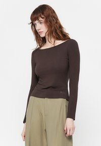Top lung cu mâneci lungi, de culoare maro, cu decolteu tip bateau, design ajustat și un detaliu mic brodat la tiv, asortat cu pantaloni de culoare verde deschis.