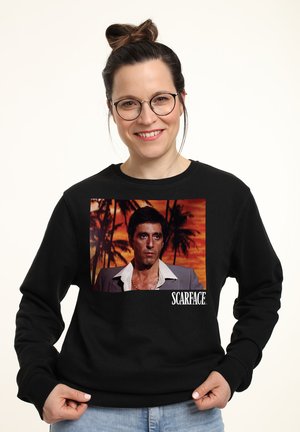Sweatshirt noir avec une photo imprimée d'un homme devant un décor de palmier, avec le texte "SCARFACE." en blanc en bas.