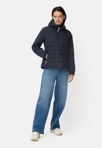 Marineblaue Steppjacke mit Kapuze, Reißverschluss und Seitentaschen; mit gesteppter Musterung und elastischen Bündchen. Kombiniert mit Jeans mit weitem Bein und Turnschuhen.