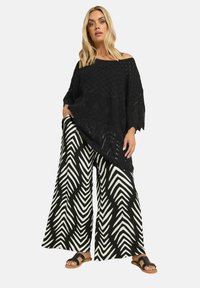 Musta virkattu poncho avoimella neuloksella, yhdistettynä leveälahkeisiin musta-valkoisiin soikean muotoisiin housuihin ja mustiin sandaaleihin.