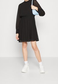 Femme portant une robe à motifs noire à manches longues, des chaussettes blanches, des baskets blanches à semelle épaisse, et tenant un sac à bandoulière bleu clair.