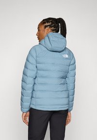 Light blue puffer jacket with a hood, featuring stitched panels and a small white logo on the upper back. Accompanied by black pants.