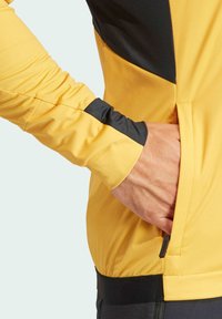 Yellow and black athletic jacket featuring a zippered pocket. The fabric appears smooth, with a fitted design and contrasting sleeve detail.