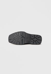 Suela de zapato de goma negra con patrones de tracción geométricos y un logo central, exhibida sobre un fondo blanco.