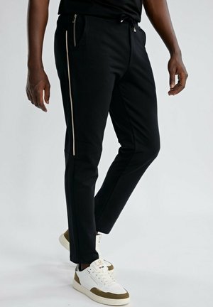 Homme portant un pantalon noir fuselé avec une fermeture éclair latérale et des baskets blanches avec des accents marron, debout devant un fond clair.