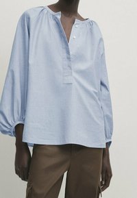 Blouse en coton bleu clair avec encolure ronde à détails froncés, patte de boutonnage à trois boutons, et manches longues larges et bouffantes.