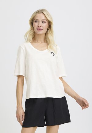 Blondína v bielom krátkodoskovom tričku s výstrihom do V a malým logom palmy, v čiernych voľných šortkách, na jednoduchom pozadí.