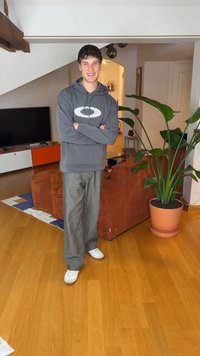 Siva jopa z logotipom, široke sive hlače in beli supergi. V ozadju je lončnica in kavč iz korduroja v svetli sobi.