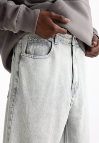 Dark-skinned person adjusting gray sweatshirt over light gray denim jeans with visible front pocket and stitching details.