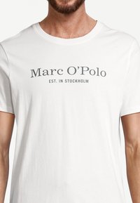 Mężczyzna nosi biały t-shirt z napisem "Marc O'Polo EST. W Sztokholmie" w szarym kolorze, widoczny od szyi do talii.