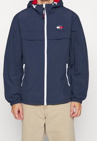 Marinblå zip-jacka med en rödfodrad huva, vit dragkedja och två sidofickor. Har en horisontell bröstficka med logotyp.