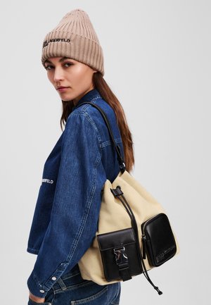 Beanie i lys beige, dongerijakke, og en beige kanvasryggsekk med svart skinnakent og glidelommer.