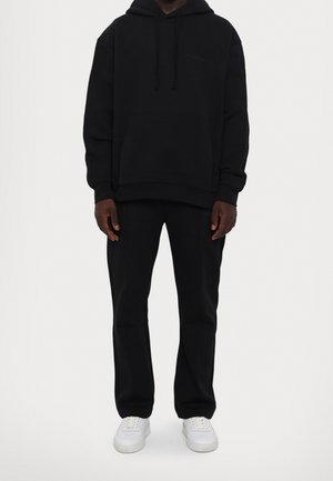Svart bomullshoodie med framficka och justerbar dragsko i huvan, tillsammans med svarta joggingbyxor och vita sneakers. Enkel, modern design.