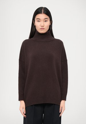 Jersey de cuello alto marrón oscuro con una textura de punto grueso, ajuste relajado y puños y dobladillo acanalados. Combinado con pantalones oscuros con pliegues.