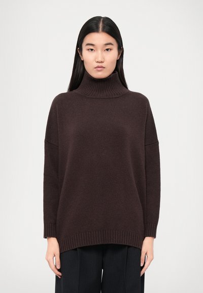 Maglione a collo alto marrone scuro con una texture a maglia spessa, vestibilità rilassata e polsini e orlo a coste. Indossato con pantaloni scuri a pieghe.