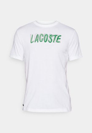 T-shirt branca de algodão com "LACOSTE" impresso em verde na parte da frente. Mangas curtas, gola redonda e pequeno logotipo na parte inferior esquerda.