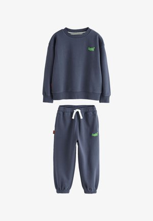 Barn med lange fletninger iført marineblå sweatshirt og bukser, holder en kasket i den ene hånd, håret blæser til siden, stående mod en ensfarvet baggrund.